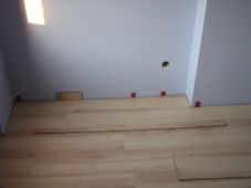 Fragment podłogi z widocznymi panelami w trakcie układania, z użyciem czerwonych klinów dystansowych przy ścianie. Widoczny kawałek niezabezpieczonej ściany i gniazdka.